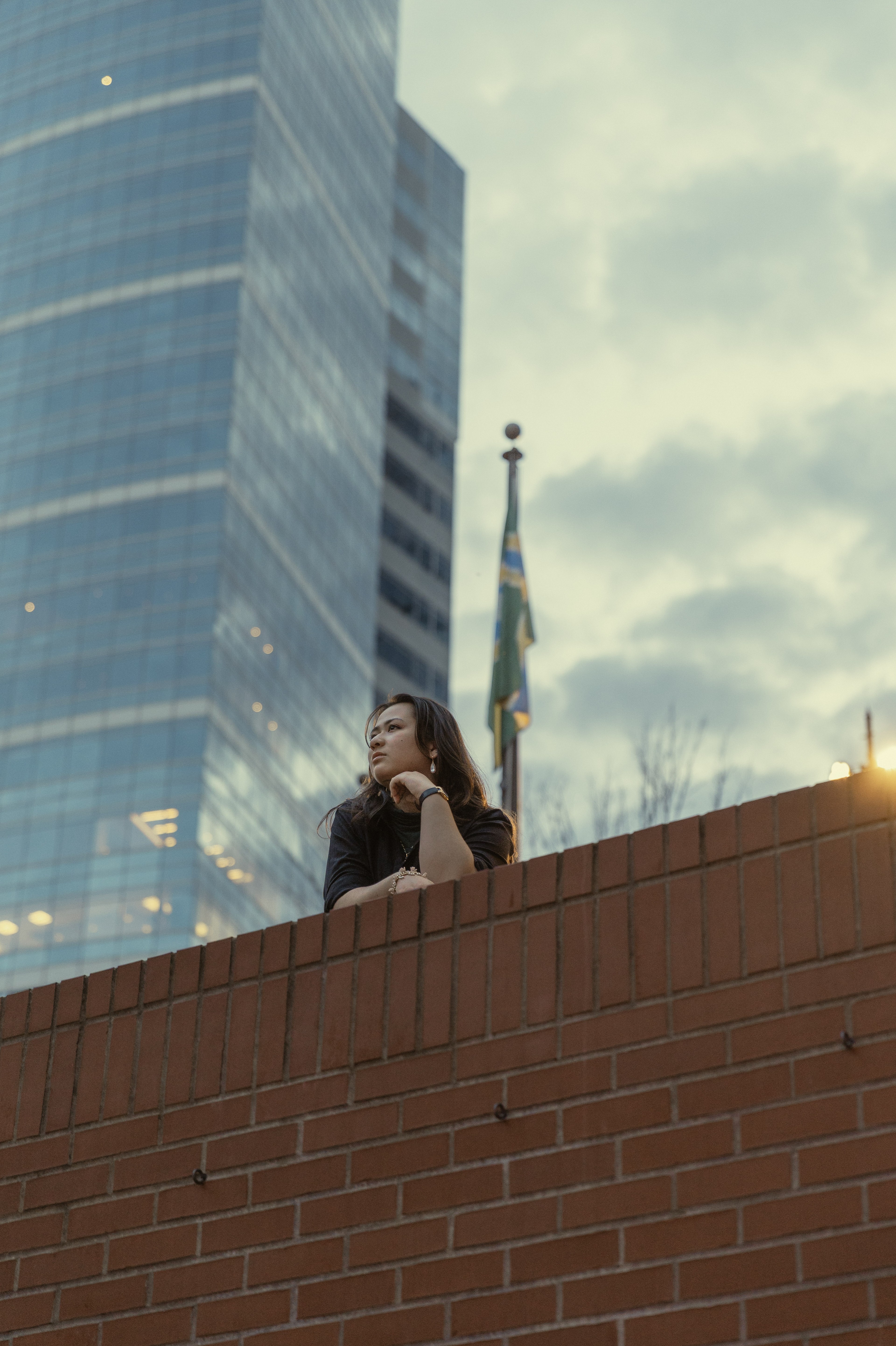 A beautiful young woman with dark hair (Simone) in a blue city scene looking up into the sky with the sunset and tall skyscrapers behind her