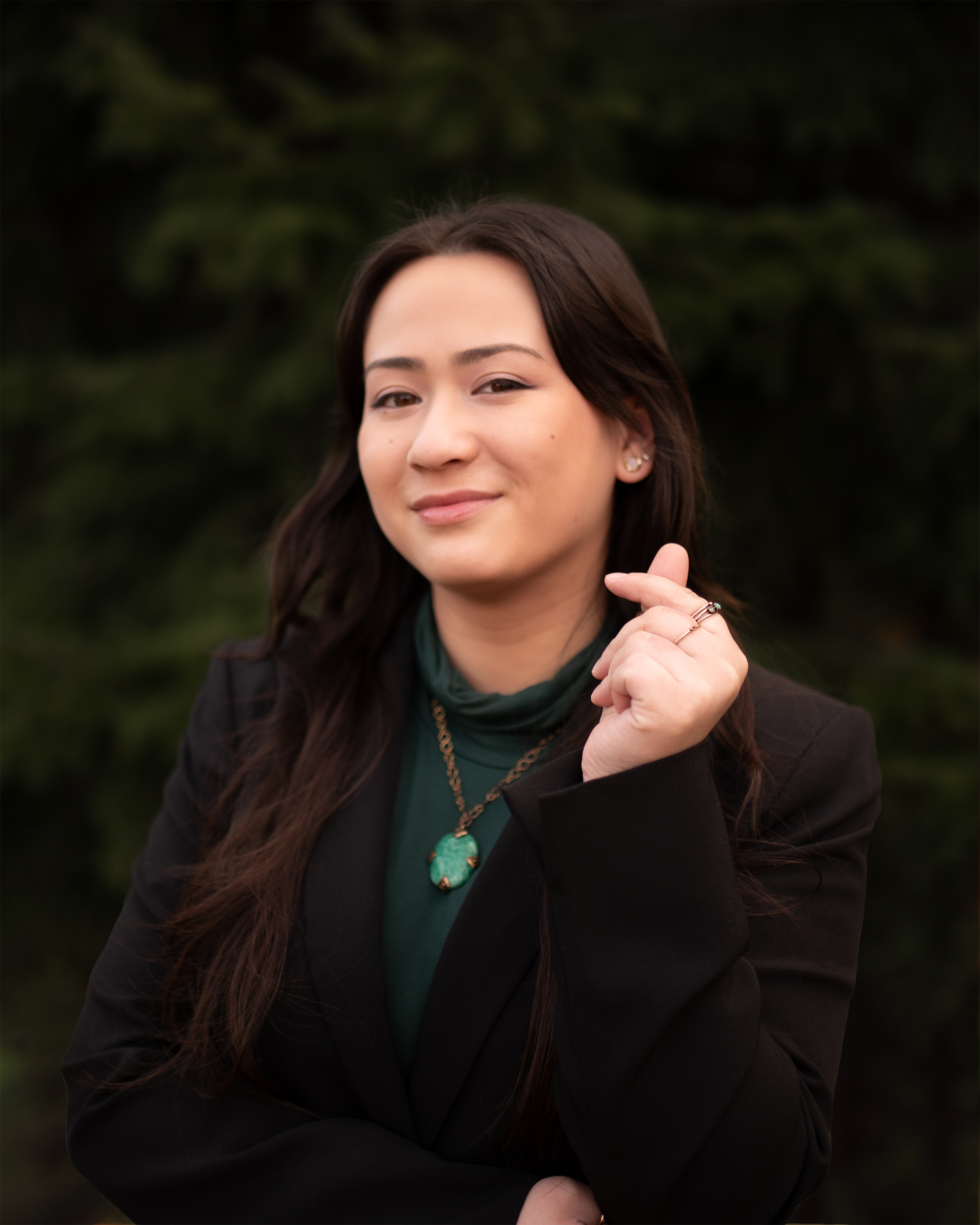 A beautiful young woman with dark hair and brown eyes (Simone) in a professional green and black outfit in front of a lush green background